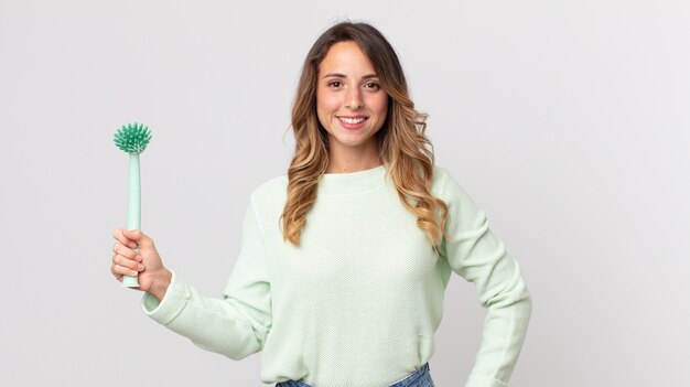 Pretty thin woman smiling happily with a hand on hip and confident and holding a washing dish brush