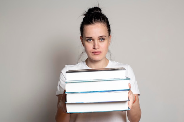 Pretty tired student woman holds a stack of books in hands and calmly stares intently at camera