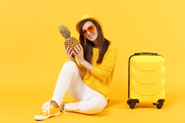 Pretty traveler tourist woman in summer clothes, hat hold fresh ripe pineapple fruit isolated on yellow orange background. Passenger traveling abroad on weekends getaway. Air flight journey concept.