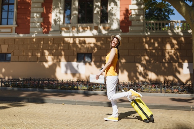 Pretty traveler tourist woman in yellow summer casual clothes hat with suitcase city map walking in city outdoor. Girl traveling abroad to travel on weekend getaway. Tourism journey lifestyle concept.