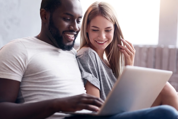 Pretty weekends. Laughing international couple using laptop and sitting together while enjoying good times.