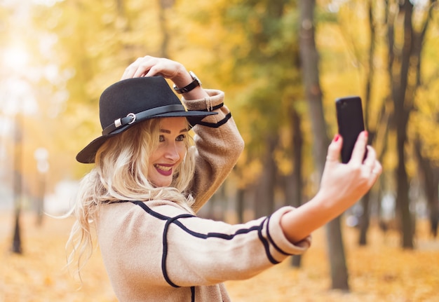 Pretty woman in black hat making selfie with her smartphone in the autumn park