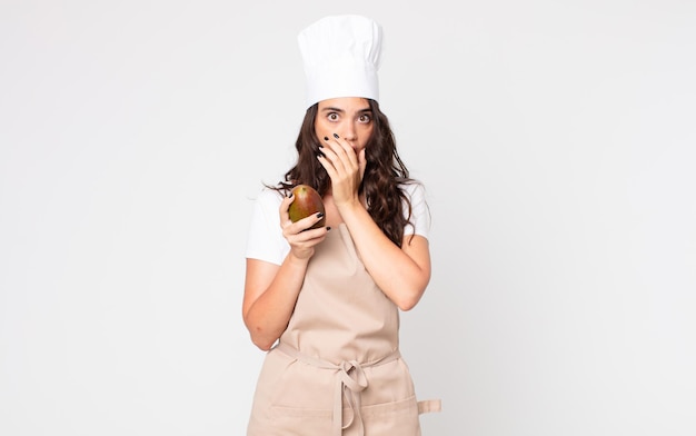 Pretty woman covering mouth with hands with a shocked wearing an apron and holding a mango