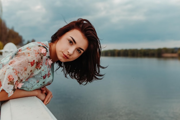 Pretty woman dressed flowery dress standing on the pier near river lake moody cloudy weather