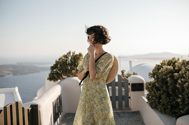 Pretty woman in floral dress smiles and enjoys sea view Happy brunette lady in summer outfit poses in Greek town with white buildings