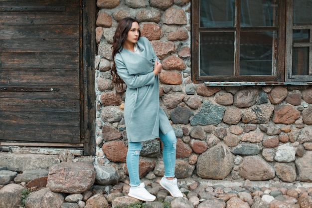 Pretty woman in a gray trendy coat, jeans and white shoes near a stone old wall