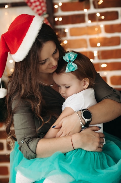 Pretty woman holding daughter in hands, standing in decorated studio with fir branches and light garland