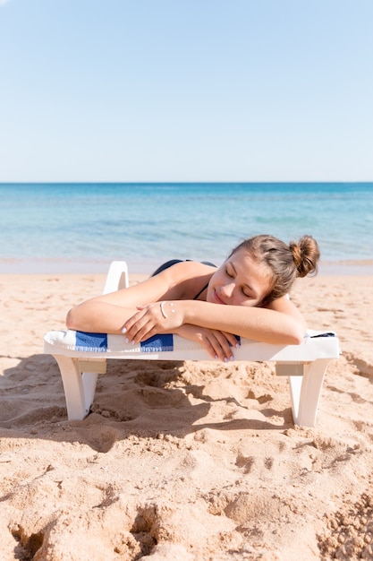 Pretty woman is sunbathing on the sunbed at the beach and has smiling face made of sunblock on her hand.