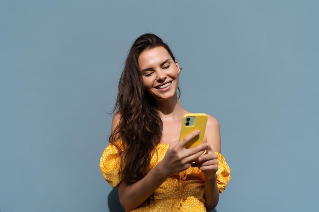 Pretty woman long dark hair in bright yellow summer dress posing on blue wall background daylight typing chatting reading messages news on mobile phone