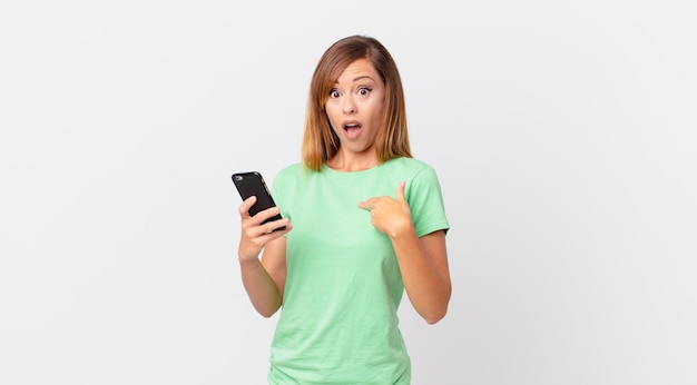 Pretty woman looking shocked and surprised with mouth wide open, pointing to self and using a smartphone