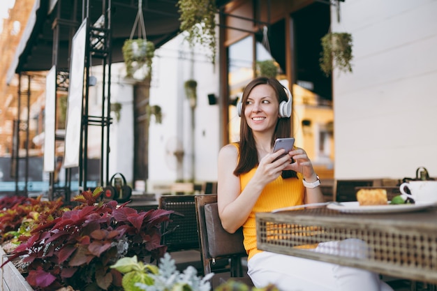 Pretty woman in outdoors street coffee shop cafe sitting at table, listen music in headphones, using mobile phone, relaxing in restaurant on free time