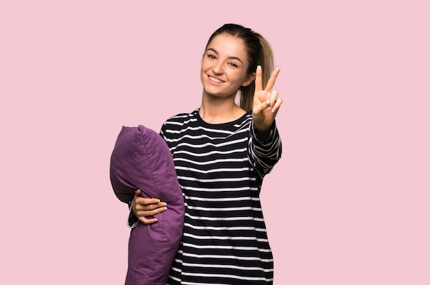 Pretty woman in pajamas smiling and showing victory sign on isolated pink wall