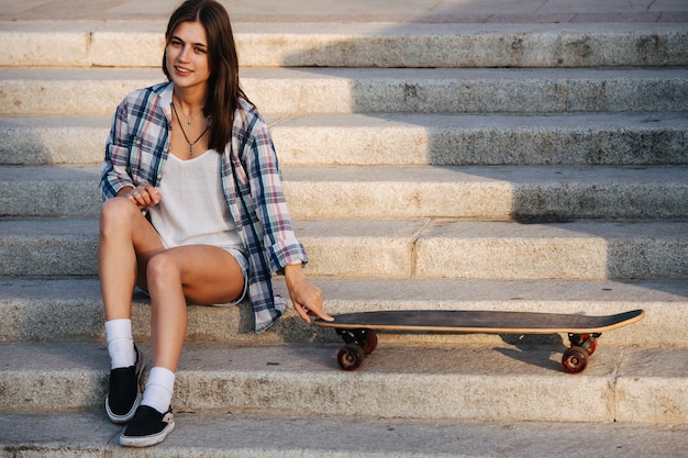 Pretty woman sitting on the stairs next to her skateboard under a gentle sun