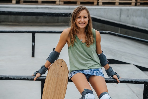 Pretty woman skater sitting on a railing at skatepark with concrete paving. Frontal view, portrait, looking at the camera.