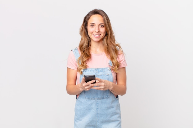 Pretty woman smiling happily with a hand on hip and confident and holding a smartphone