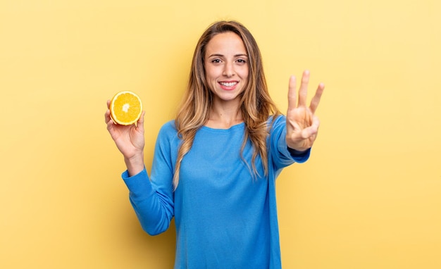 Pretty woman smiling and looking friendly, showing number three holding half orange