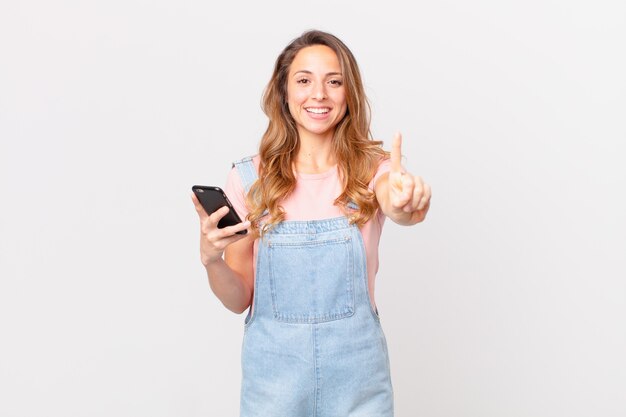 Pretty woman smiling proudly and confidently making number one and holding a smartphone