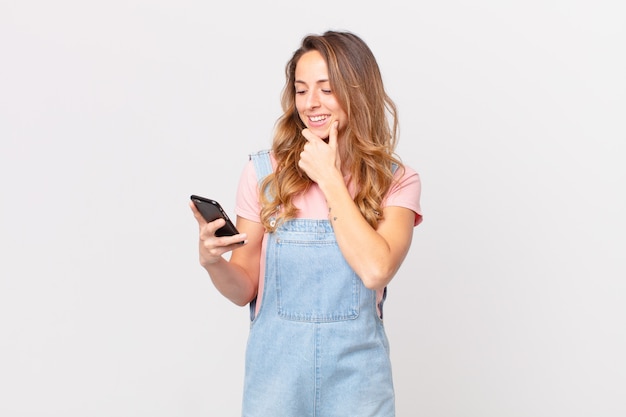 Pretty woman smiling with a happy, confident expression with hand on chin and holding a smartphone