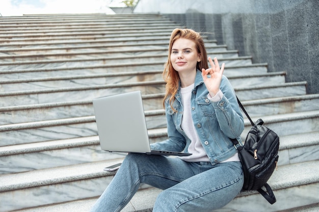 Pretty woman student with laptop sits on the stairs outdoors Distance education