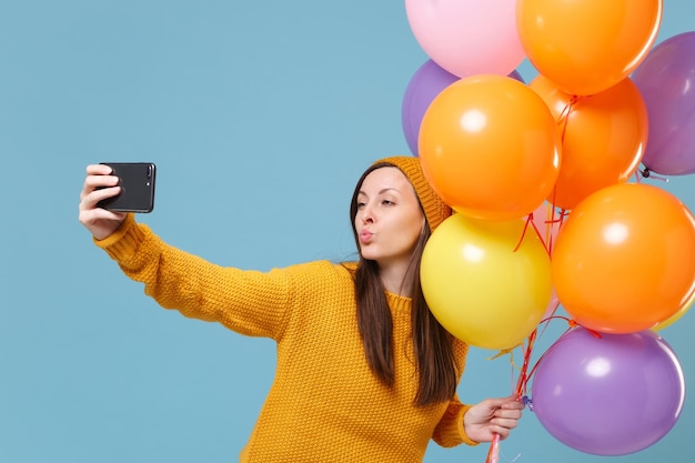 Pretty woman in sweater hat posing isolated on blue background. Birthday holiday party, people emotions concept. Mock up copy space. Celebrating hold air balloons doing selfie shot on mobile phone.