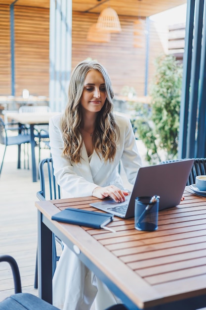 A pretty woman in a white classic suit is working with a laptop on an open terrace in a cafe A woman in stylish business clothes works remotely on a laptop in a summer cafe