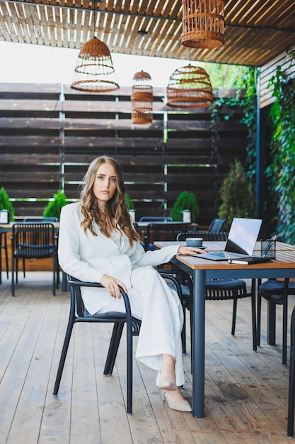 A pretty woman in a white classic suit is working with a laptop on an open terrace in a cafe A woman in stylish business clothes works remotely on a laptop in a summer cafe