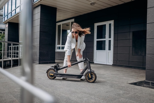 Pretty woman in white suit folding her electro scooter after ride while standing on background of modern building