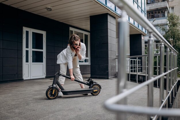 Pretty woman in white suit folding her electro scooter after ride while standing on background of modern building