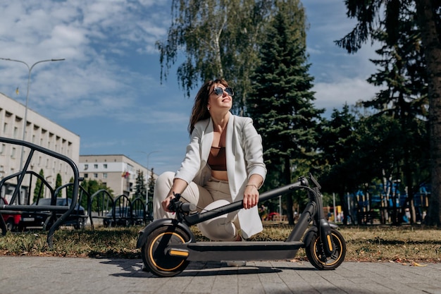 Pretty woman in white suit folding her electro scooter after ride while standing in city parkland