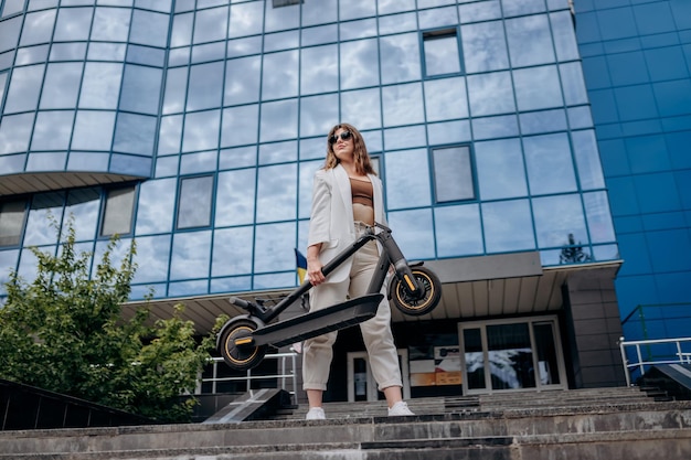 Pretty woman in white suit holding her electro scooter after ride while standing on background of modern building