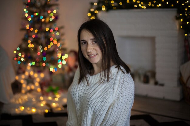 A pretty woman in white sweater smiling and in front of christmas tree and fireplace.