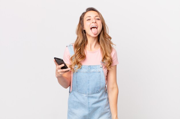 Pretty woman with cheerful and rebellious attitude, joking and sticking tongue out and holding a smartphone