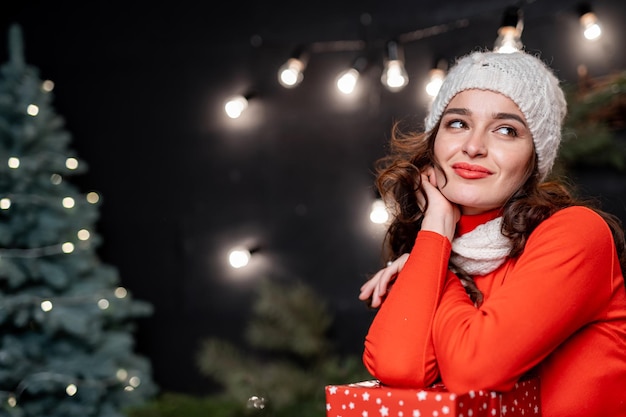 Pretty woman with hand near face poses to camera while looking away dreamy. Red sweater on a nice girl. New year decorations. Holidays concept