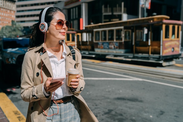 Pretty woman with headphones and cellphone in city streets. young elegant smiling lady holding cup of hot coffee beverage walking on sunlight. cable car on san francisco road usa in background.