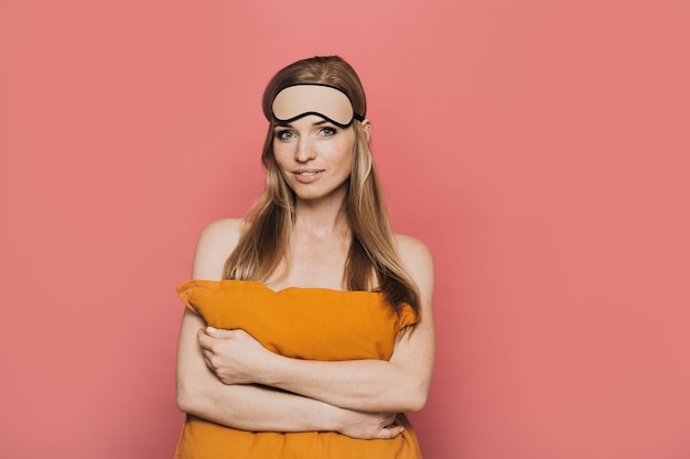 Pretty woman with mask for sleep on her head, smiling tenderly, hugging an orange pillow, looking satisfied, over pink background.