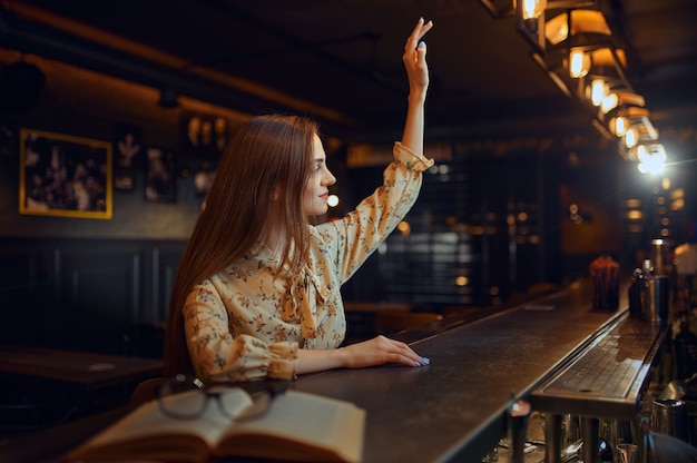 Pretty woman with raised hand sitting at the counter in bar. One female person in pub, human emotions, leisure activities, nightlife