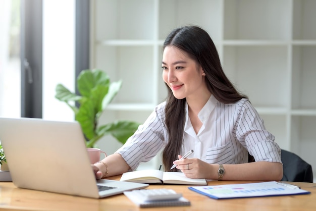 Pretty young Asian businesswoman working on laptop and taking notes In office.