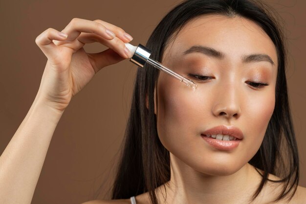 Pretty young asian lady using nourishing face serum after shower enjoying organic beauty product brown background