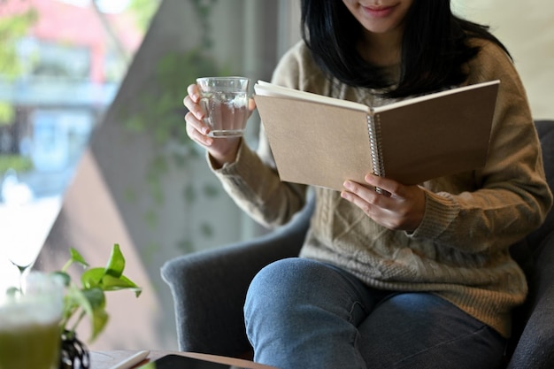 Pretty young Asian woman reading a book sipping water while relaxing in the coffee shop