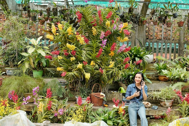 Pretty young Asian woman working in greenhouse and talking on phone when ordering new plants for selling