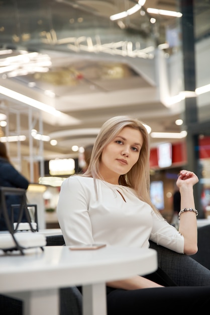 Pretty young beautiful blonde woman with long hair, sitting in cafe in mall. Attractive modern girl in trendy clothes is enjoys hot drink indoors.