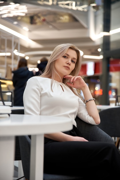 Pretty young beautiful blonde woman with long hair, sitting in cafe in mall. Attractive modern girl in trendy clothes is enjoys hot drink indoors.