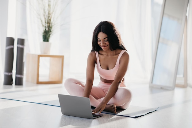 Pretty young black woman in sports clothes using laptop sitting on mat in lotus pose speaking to her