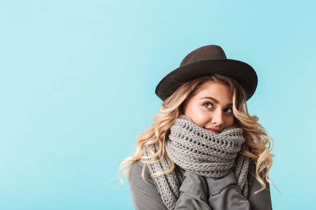 Pretty young blonde woman wearing hat and scarf looking away at copy space isolated over blue wall