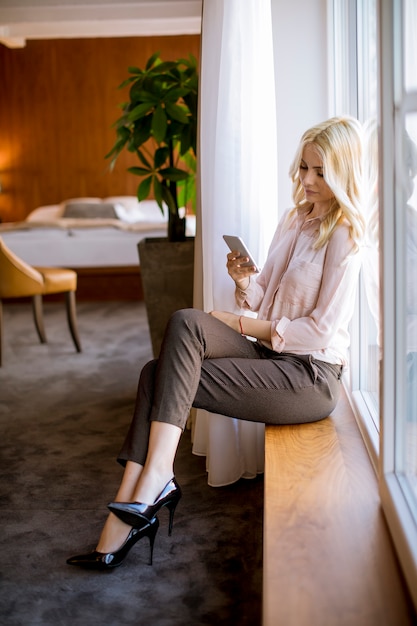 Pretty young blonde woman with mobile phone by the window in the room