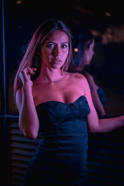 A pretty young brunette Caucasian woman in a black dress illuminated by purple LED lights, looking at the camera. Night urban photography