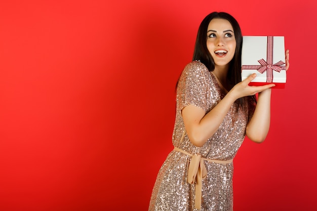 Pretty young brunette woman dressed in beige dress shakes gift box to find out what's inside.