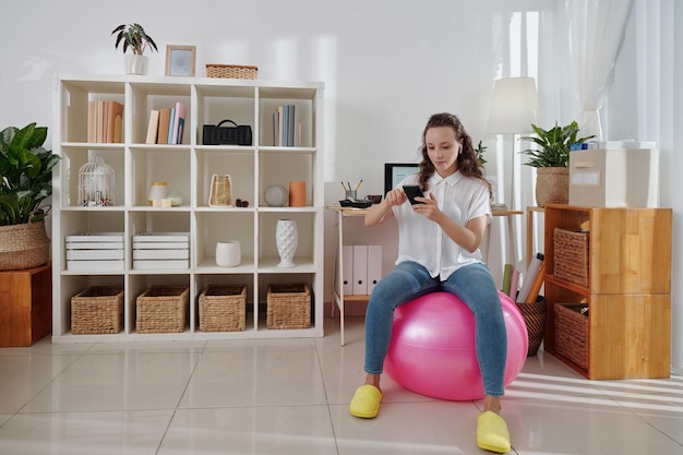 Pretty young businesswoman sitting on fitness ball and answering text messages from ccolleagues on smartphone