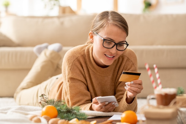 Pretty young casual woman with smartphone and credit card looking at personal data while going to pay for online order