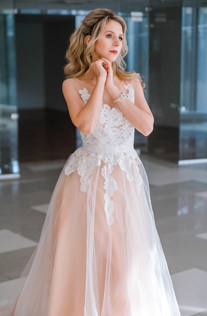 Pretty young caucasian blonde bride with makeup and trendy hairstyle in wedding dress standing and posing in interior